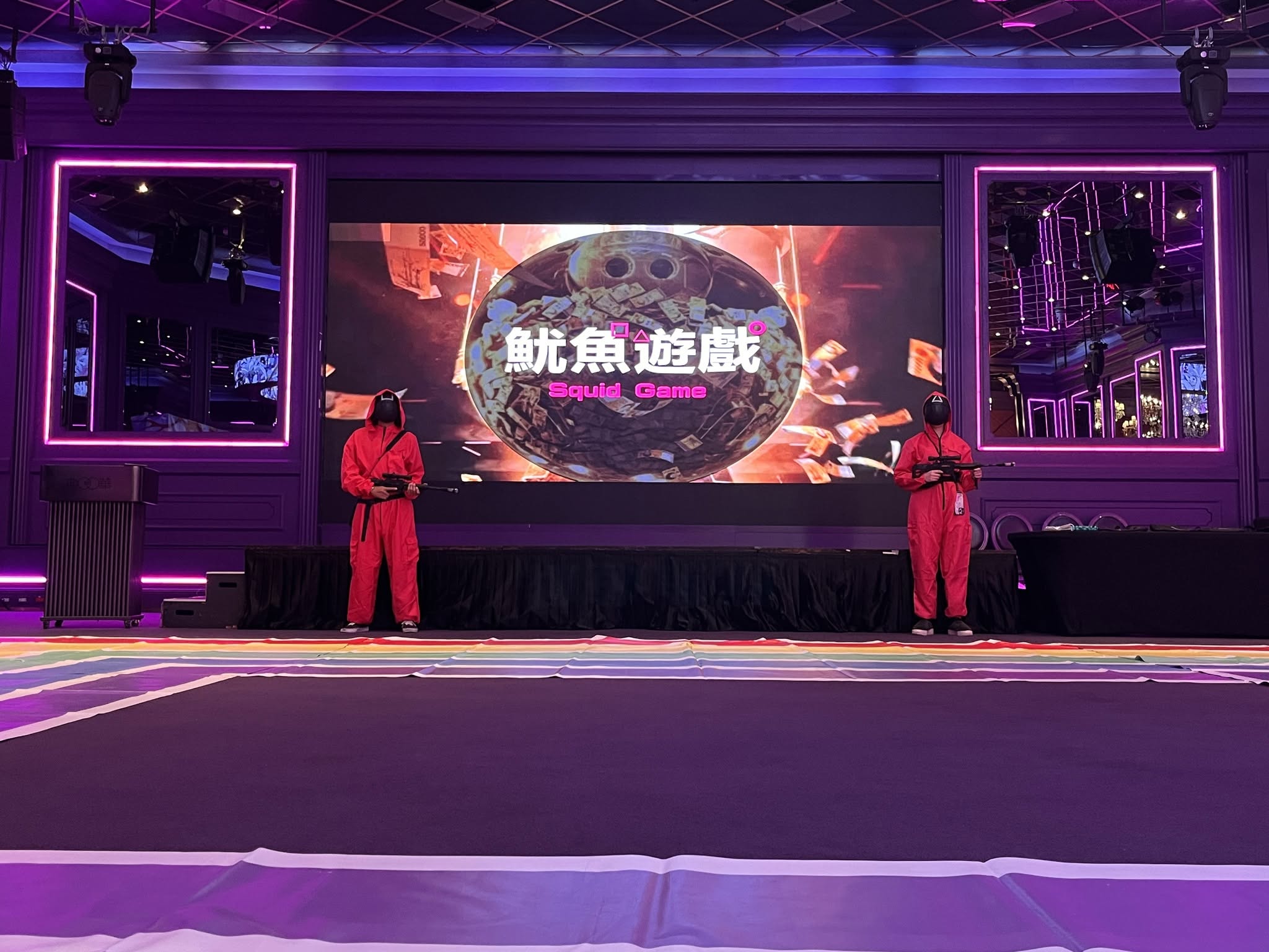 瘋戶外企業 Team Building 魷魚遊戲真人版活動現場。紫色霓虹燈光的氣派舞台上，大螢幕投影著「魷魚遊戲 Squid Game」經典字樣與滿滿金幣的巨型撲滿，兩側有身穿紅色工作服、配戴幾何圖形面具的工作人員持道具守衛，營造高沉浸感的活動氛圍。